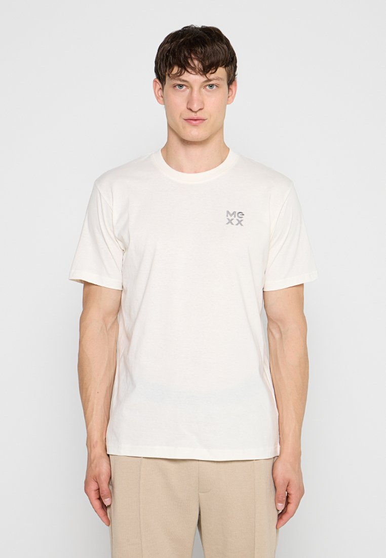 Junger Mann mit kurzen braunen Haaren, der ein schlichtes weißes T-Shirt mit dem Schriftzug „MCXX“ auf der Brust und beigefarbene Hosen vor einem weißen Hintergrund trägt.