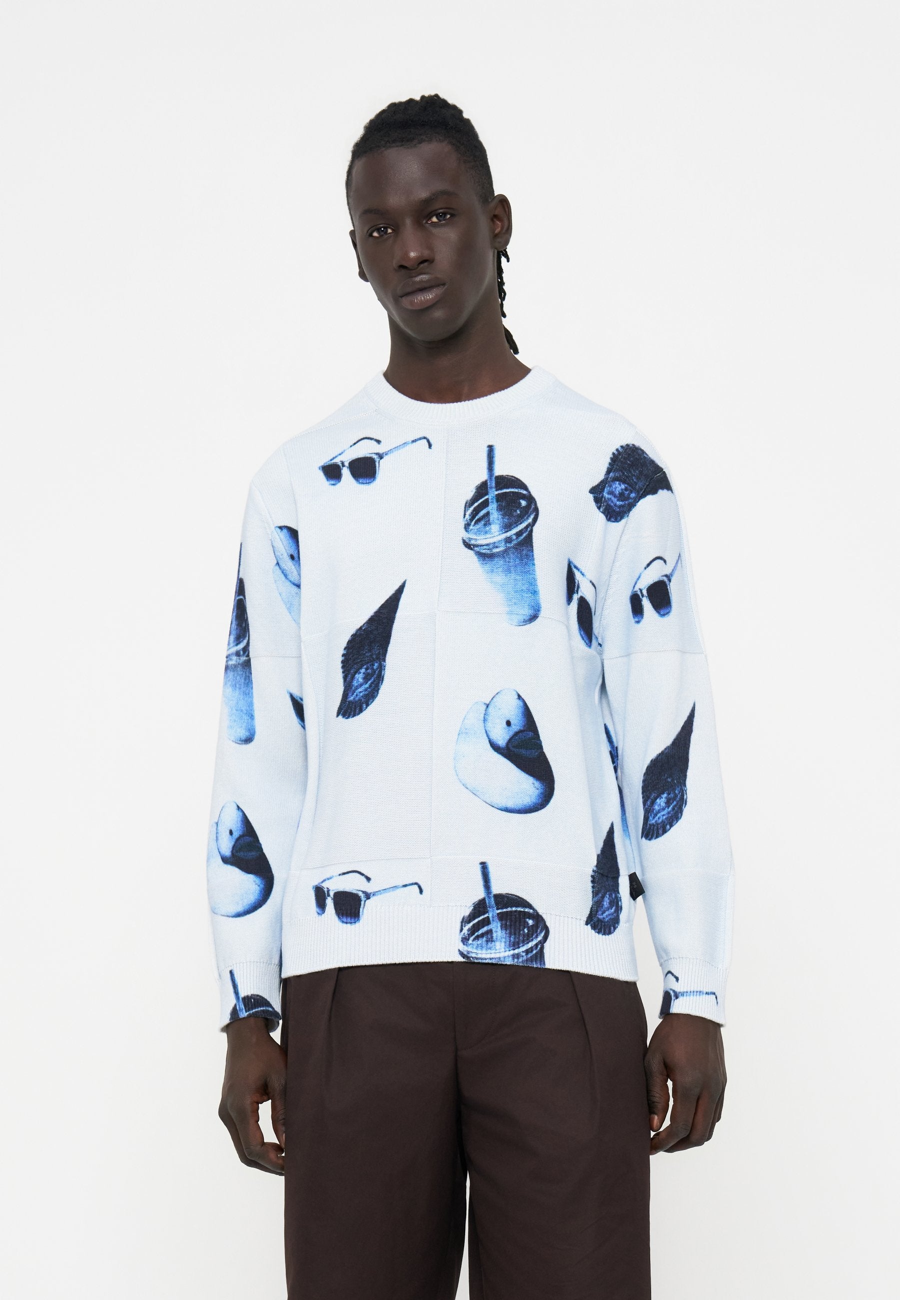 Junger Mann, der einen weißen Pullover mit blauen Aufdrucken von Sonnenbrillen, Enten und Getränken trägt, kombiniert mit dunkelbraunen Hosen vor einem einfarbigen Hintergrund.