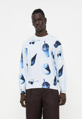 Junger Mann, der einen weißen Pullover mit blauen Aufdrucken von Sonnenbrillen, Enten und Getränken trägt, kombiniert mit dunkelbraunen Hosen vor einem einfarbigen Hintergrund.