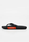Schwarze Flipflop-Sandale mit einem roten Etikett an der Seite, auf dem "BORN FROM WATER" steht, vor weißem Hintergrund.