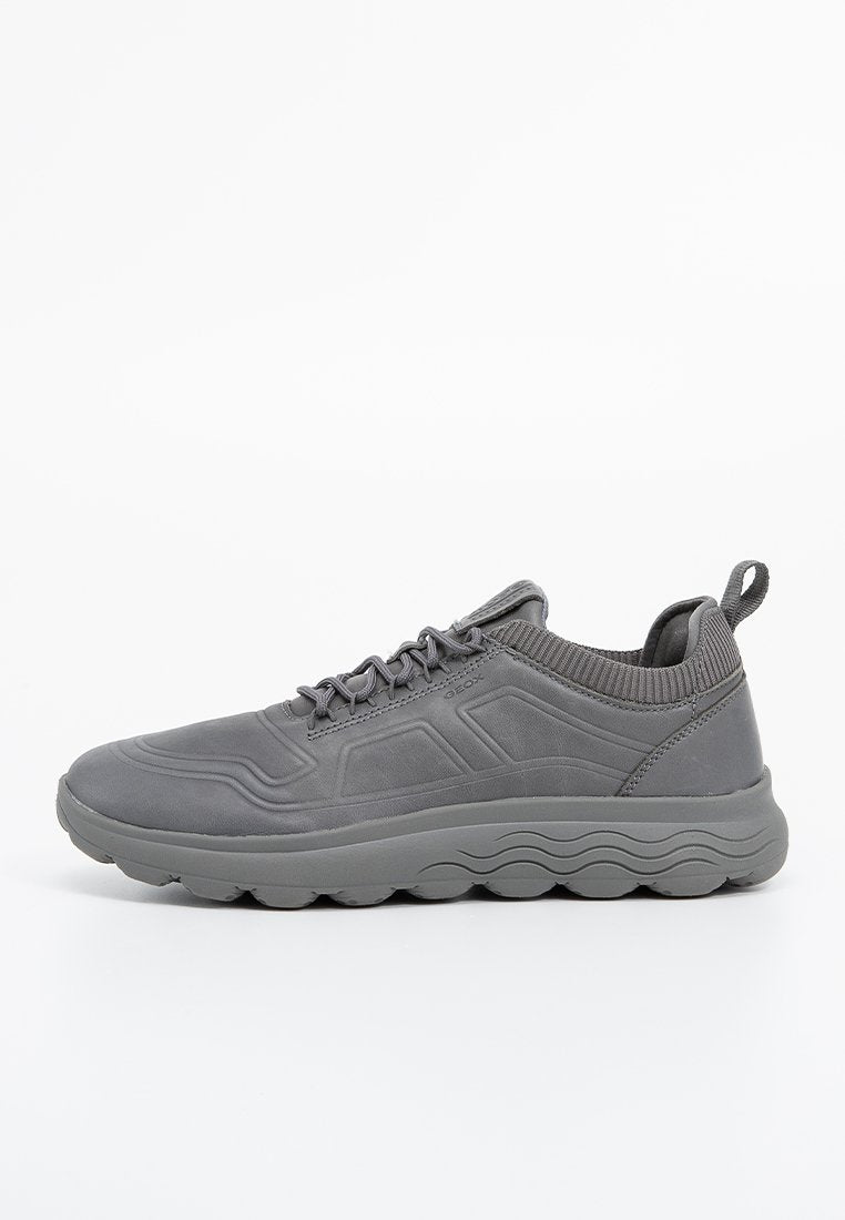 Grauer Sportschuh aus Synthetikmaterial, mit strukturiertem Obermaterial, gepolstertem Kragen und einer dicken, wellenmusterförmigen Sohle.