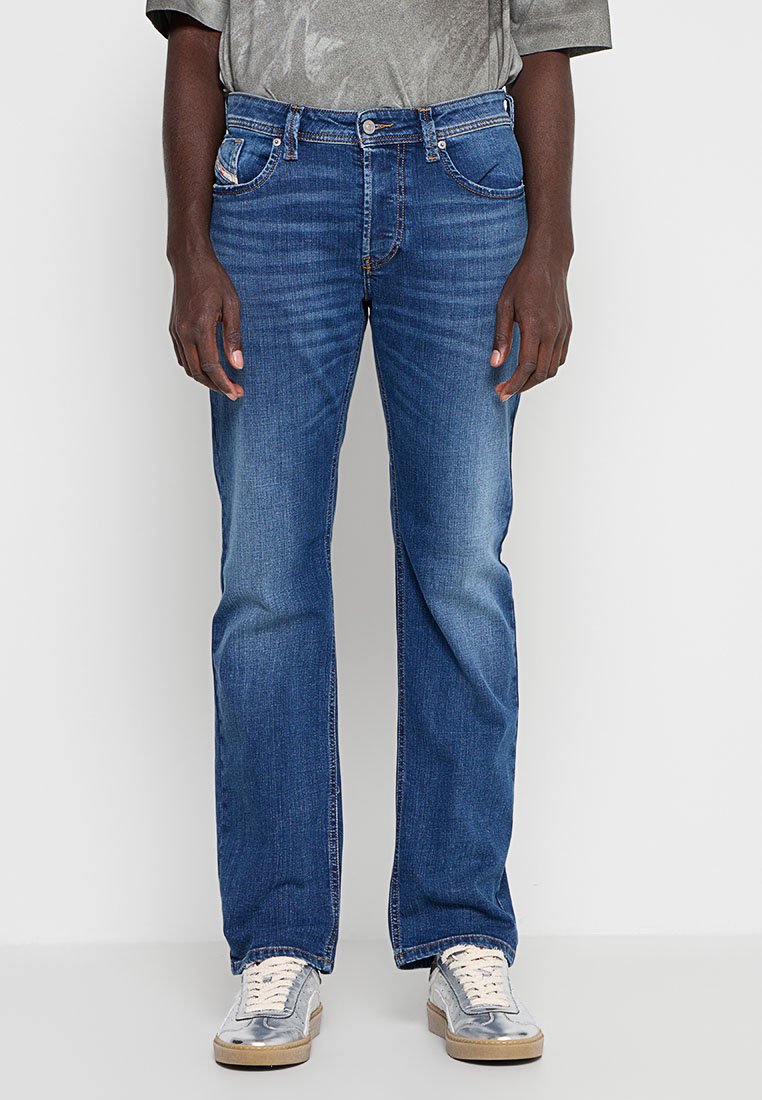 Mann, der mittelblaue, gerade geschnittene Jeans und silberne Sneakers trägt, steht vor einem schlichten, hellen Hintergrund.