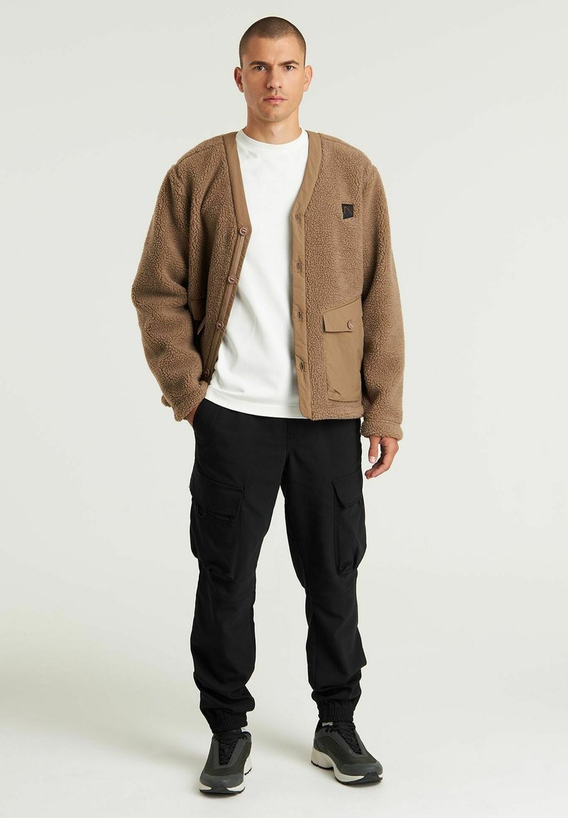 Brauner Fleece-Cardigan mit großen Taschen, Frontknöpfen und einem kontrastierenden hellbraunen Kragen, getragen über einem weißen T-Shirt und einer schwarzen Cargo-Hose.