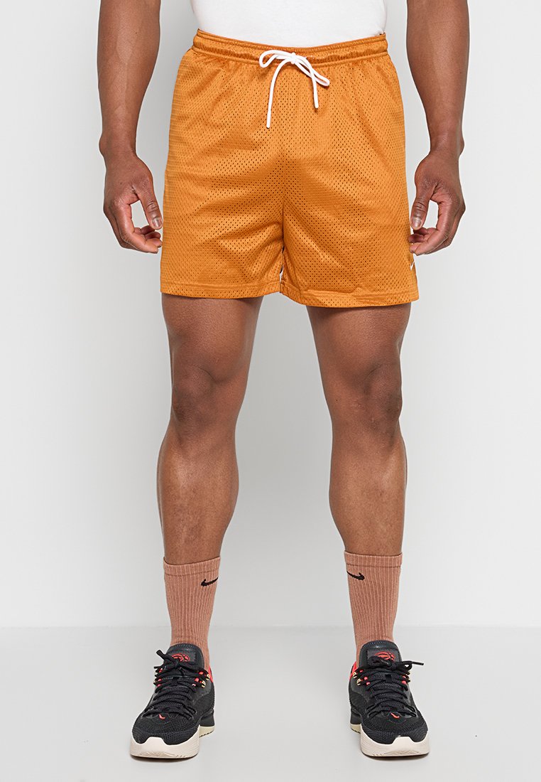 Mann trägt orangefarbene Netz-Sportshorts mit weißem Kordelzug, braune Sportsocken und schwarze Turnschuhe, steht vor einem weißen Hintergrund.
