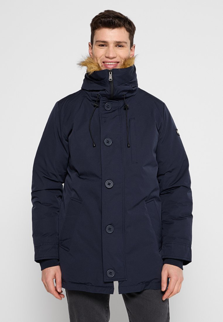 Lächelnder Mann, der eine marineblaue Winterparka mit einer fellbesetzten Kapuze, Knopf- und Reißverschluss sowie vorderen Taschen trägt, vor einem schlichten Hintergrund.