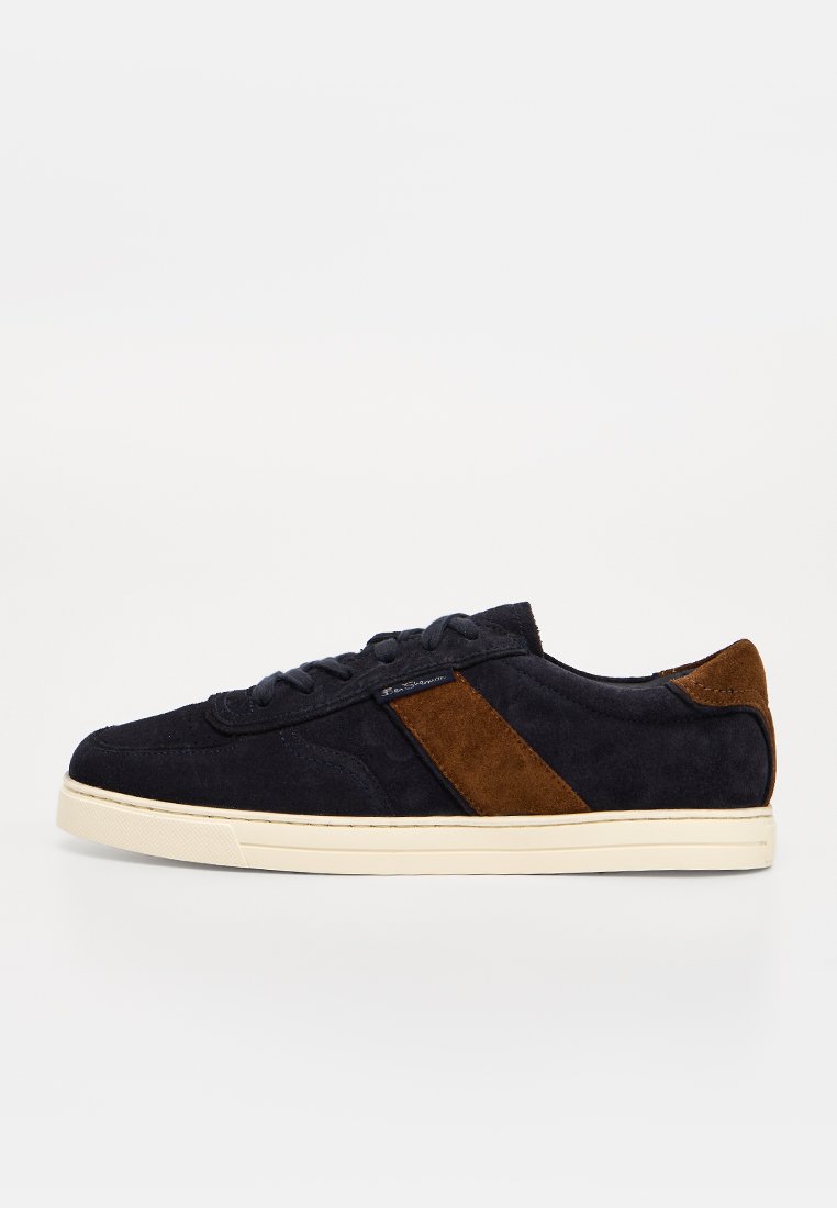 Schwarzer Veloursleder-Sneaker in niedriger Ausführung mit braunem Veloursstreifen und Fersendetail, cremefarbener Gummisohle und schwarzen Schnürsenkeln auf weißem Hintergrund.