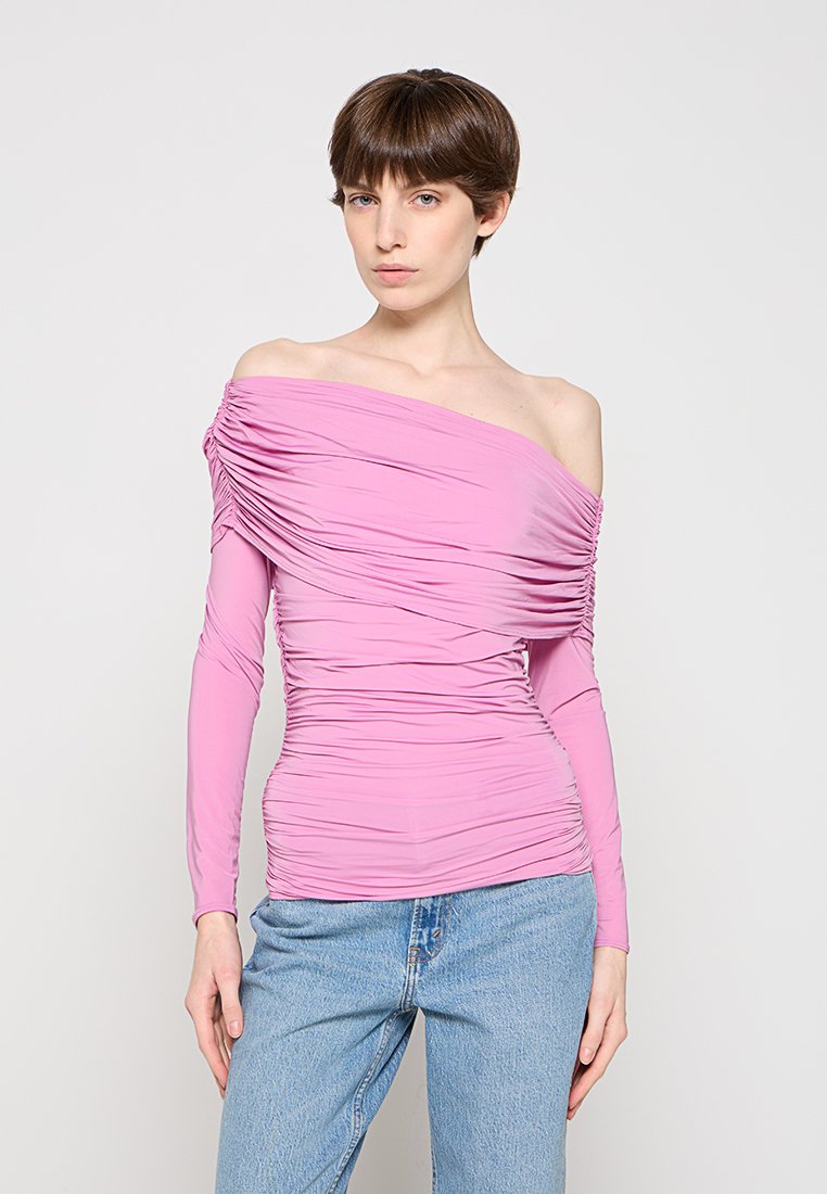 Frau mit kurzen braunen Haaren, die ein gerafftes, schulterfreies rosa Langarmshirt trägt, kombiniert mit hellblauen Jeans vor einem schlichten Hintergrund.