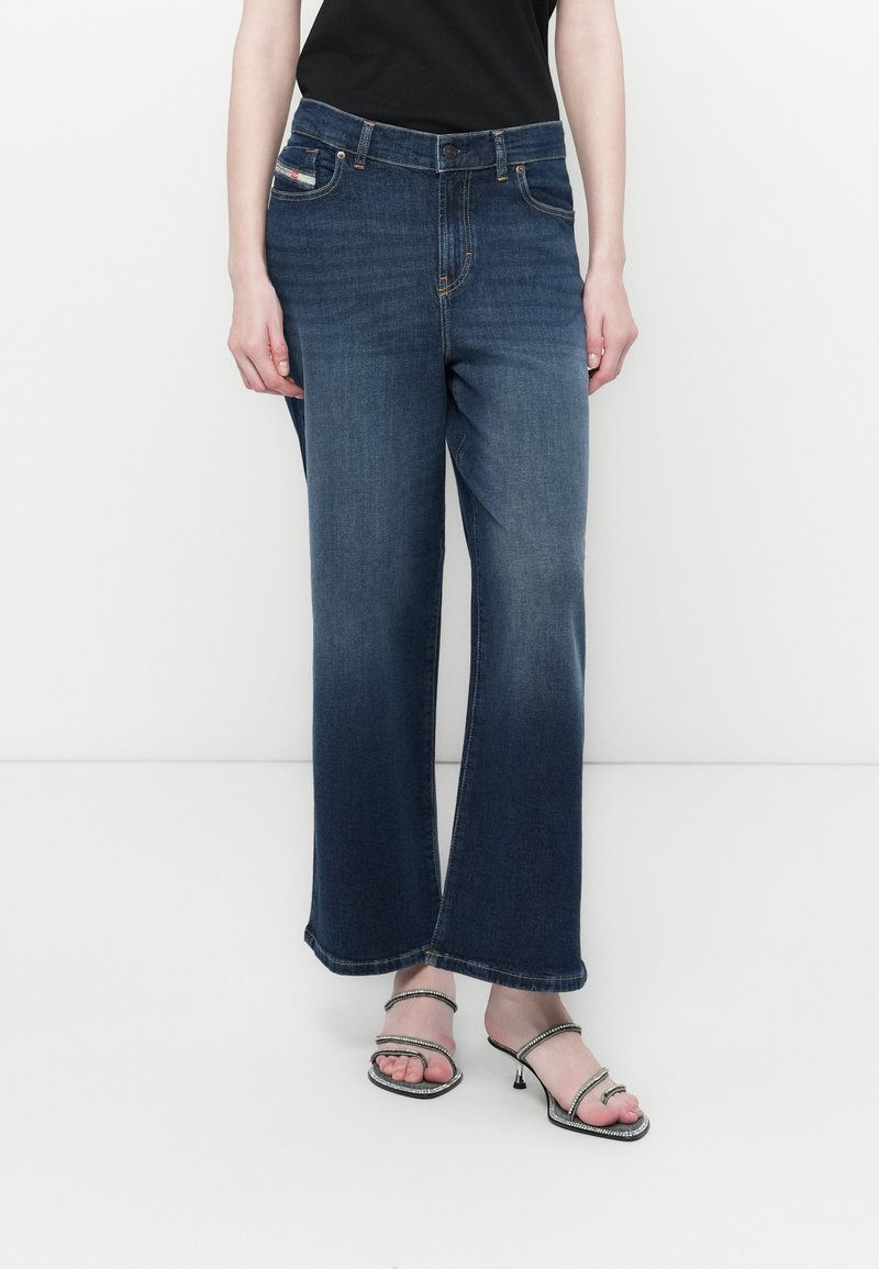 Frau in dunkelblauen, ausgestellten Jeans, schwarzem Oberteil und offenen schwarzen High Heels mit strassbesetzten Riemen, die vor einem weißen Hintergrund steht.