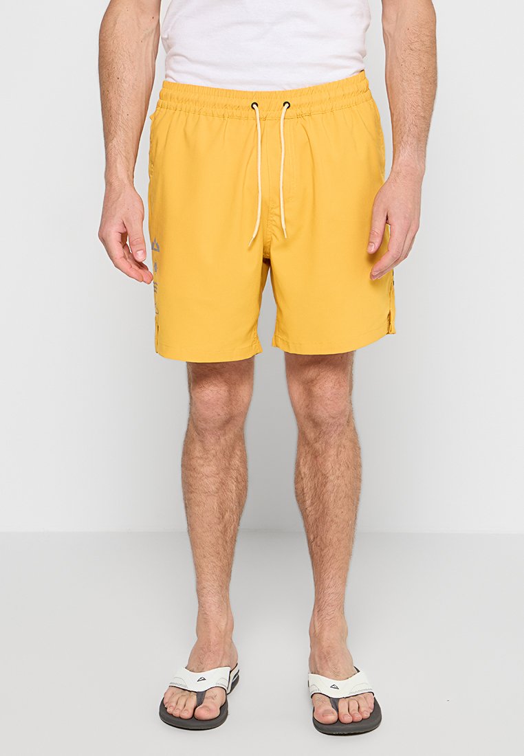 Mann in gelben Kordelzugshorts, weißem T-Shirt und weißen Sandalen, der vor einem schlichten weißen Hintergrund steht.