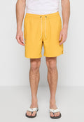 Mann in gelben Kordelzugshorts, weißem T-Shirt und weißen Sandalen, der vor einem schlichten weißen Hintergrund steht.