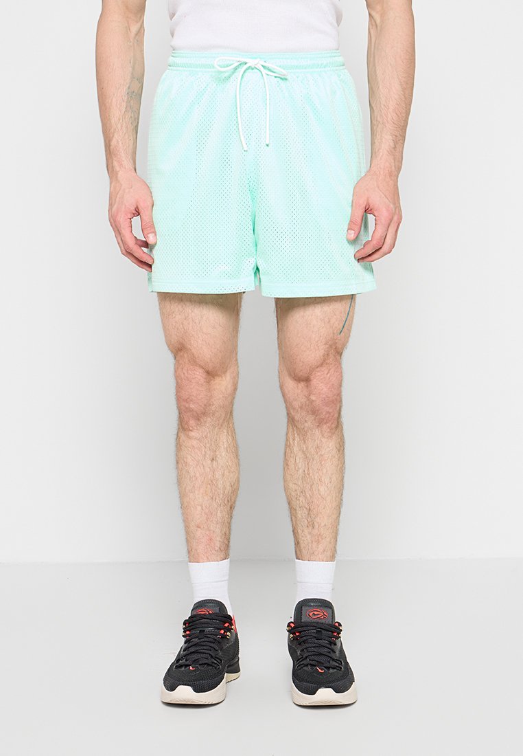 Mann trägt mintgrüne Mesh-Shorts, weiße Socken und schwarze Sportschuhe und steht vor einer weißen Wand.