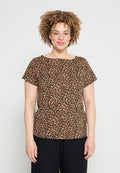 Frau mit lockigem Haar, die ein kurzärmeliges Leopardenmuster-Top und schwarze Hosen trägt, steht vor einem schlichten, hellen Hintergrund.