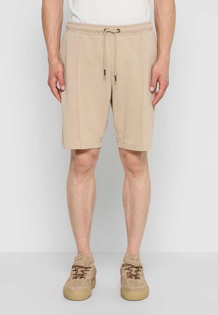 Beige Kordelzug-Shorts, getragen von einer stehenden Person, kombiniert mit beigen Sneakers mit dicken Sohlen, vor einfachem hellgrauem Hintergrund.
