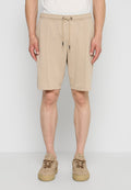 Beige Kordelzug-Shorts, getragen von einer stehenden Person, kombiniert mit beigen Sneakers mit dicken Sohlen, vor einfachem hellgrauem Hintergrund.