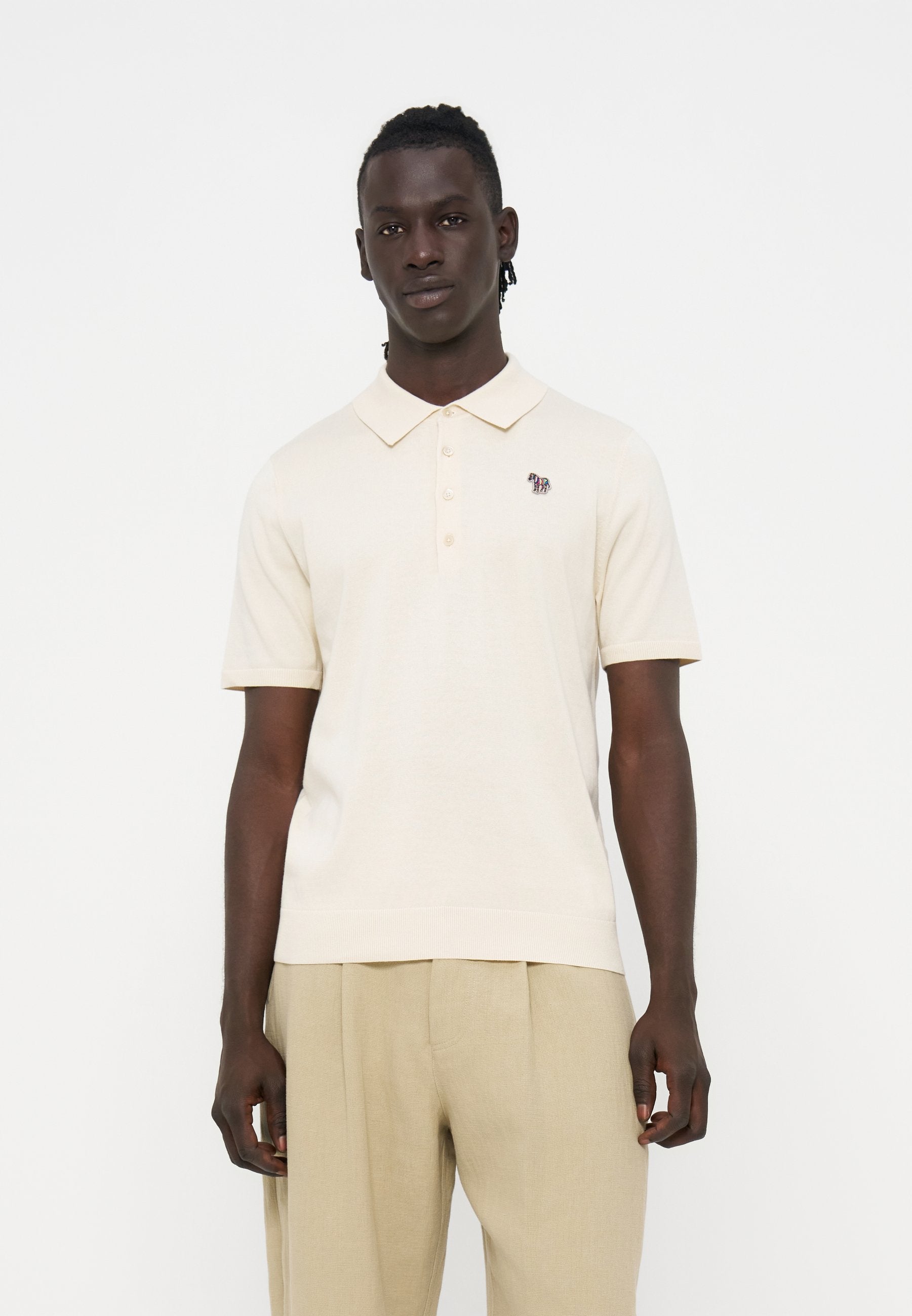 Junger Mann trägt ein cremefarbenes kurzärmeliges Poloshirt mit einem kleinen gestickten Zebra auf der Brust und hellbeigen Hosen.