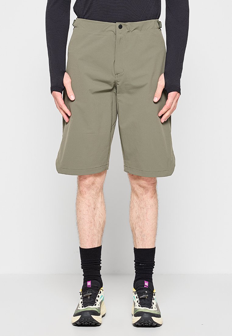Mann trägt olivgrüne knielange Shorts, schwarzes Langarmshirt, schwarze Socken und mehrfarbige Laufschuhe, steht in Frontalansicht.