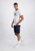 Mann trägt ein weißes Alpha Industries T-Shirt, marineblaue Cargoshorts, weiße Socken und weiße Turnschuhe, steht mit einer Hand in der Tasche.