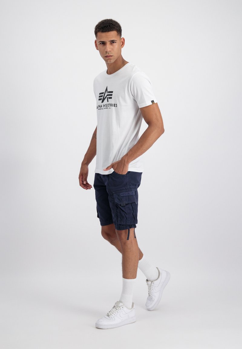 Mann trägt ein weißes Alpha Industries T-Shirt, marineblaue Cargoshorts, weiße Socken und weiße Turnschuhe, steht mit einer Hand in der Tasche.