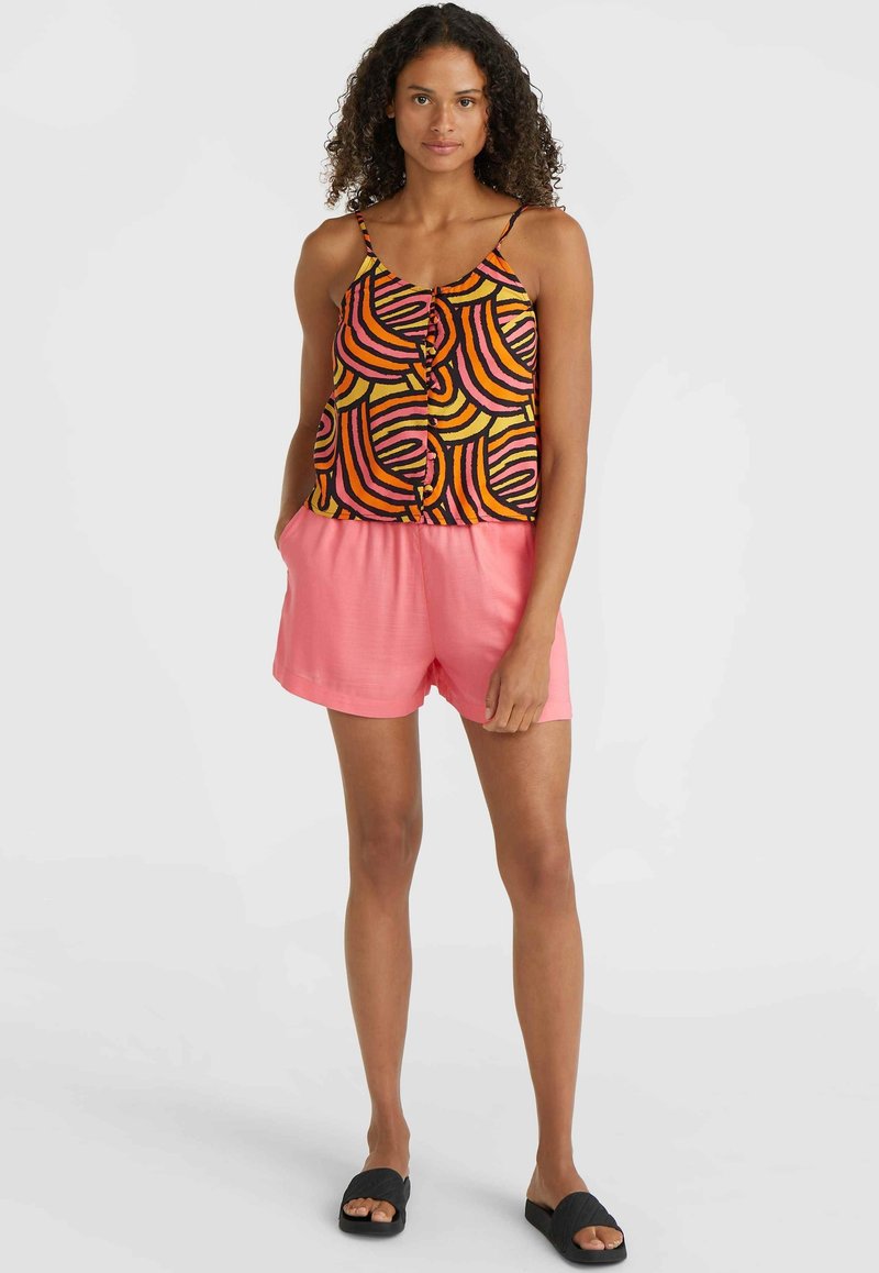 Ein mehrfarbiges, gemustertes Tanktop mit schwarzen, orangen und pinken Wellen; kombiniert mit einfarbigen pinken Shorts; das Model trägt schwarze Riemchensandalen.