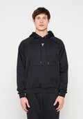 Mann steht frontal, trägt einen schwarzen Hoodie mit einem kleinen weißen Logo auf der Brust und schwarze Hosen vor einem einfarbigen hellen Hintergrund.