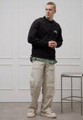 Schwarzer Pullover mit grünem Logo, darüber getragen ein grün gestreiftes Hemd. Beigefarbene Cargohose mit Taschen und helle Sneaker. Einfaches Design.