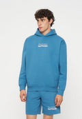 Junger Mann mit lockigem Haar, der einen blauen Hoodie und passend dazu Shorts mit weißem Text „The Future Surf Society“ auf beiden trägt.