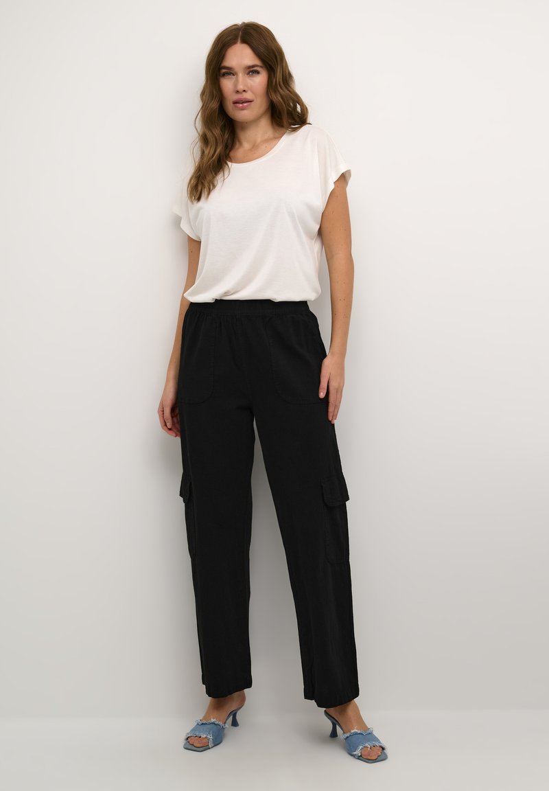 Frau mit welligem braunem Haar, die eine weiße kurzärmlige Bluse, schwarze Cargo-Hosen mit hohem Bund und blaue Peeptoe-Sandalen mit Absatz trägt.