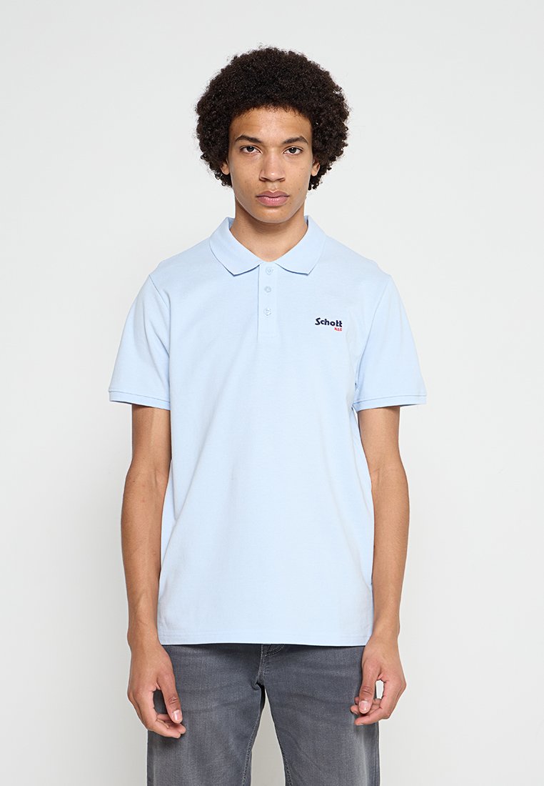 Junger Mann mit lockigem Haar, der ein hellblaues Schott Polohemd und graue Jeans trägt, steht vor einem einfarbigen Hintergrund.