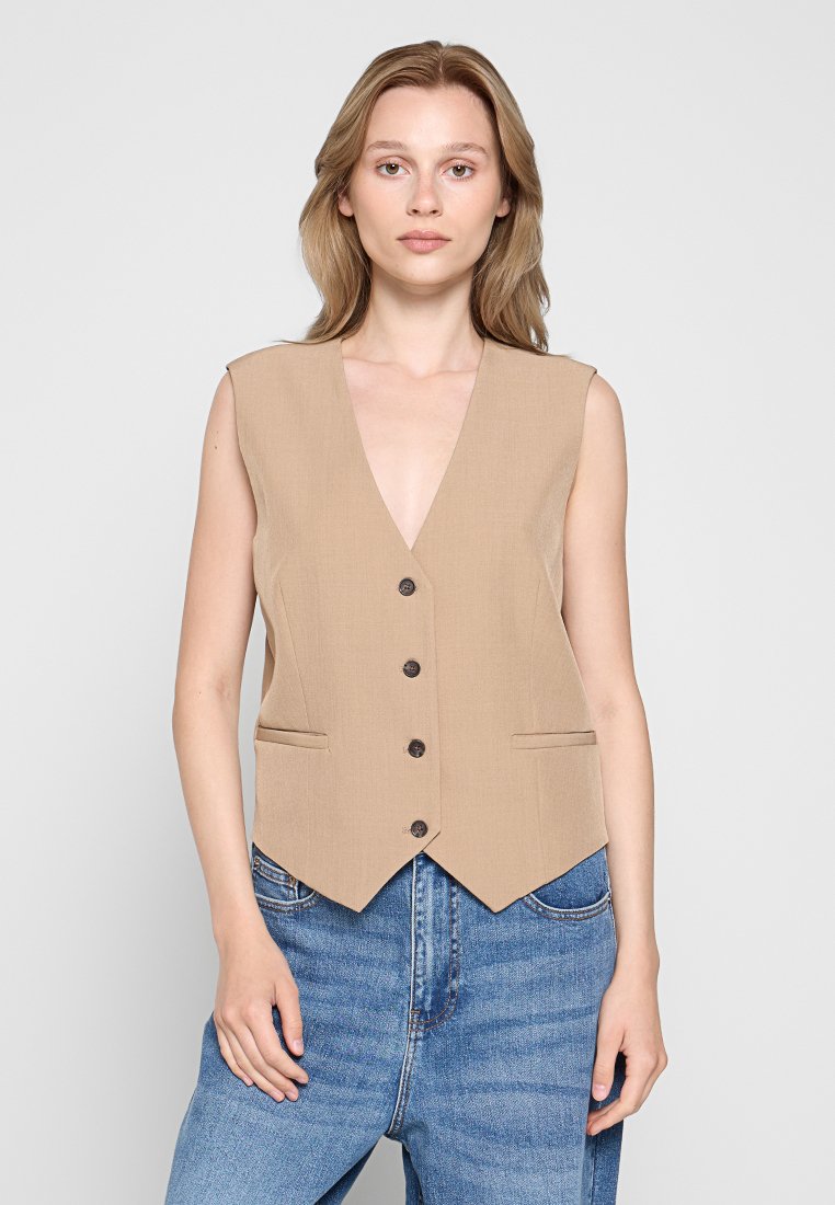 Beige ärmellose Weste mit V-Ausschnitt, fünf dunklen Knöpfen und Seitentaschen; strukturierter Stoff; getragen über blauen, hochtaillierten Jeans.