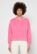 Frau mit lockigem Haar, die einen leuchtend pinken Pullover mit langen Ärmeln und hellbeige Hosen trägt, steht vor einem schlichten hellgrauen Hintergrund.