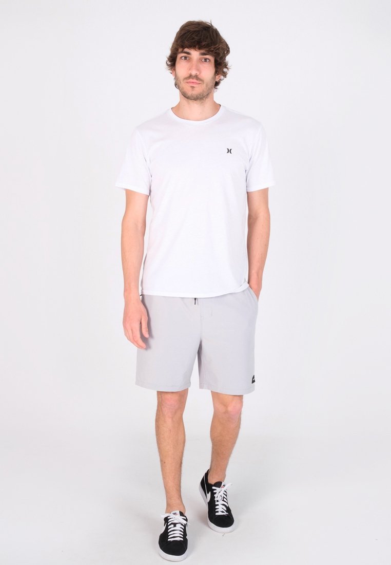 Weißes Baumwoll-T-Shirt mit kleinem schwarzem Logo, kombiniert mit hellgrauen Shorts mit Kordelzug und Seitentaschen, getragen mit schwarzen Sneakers.