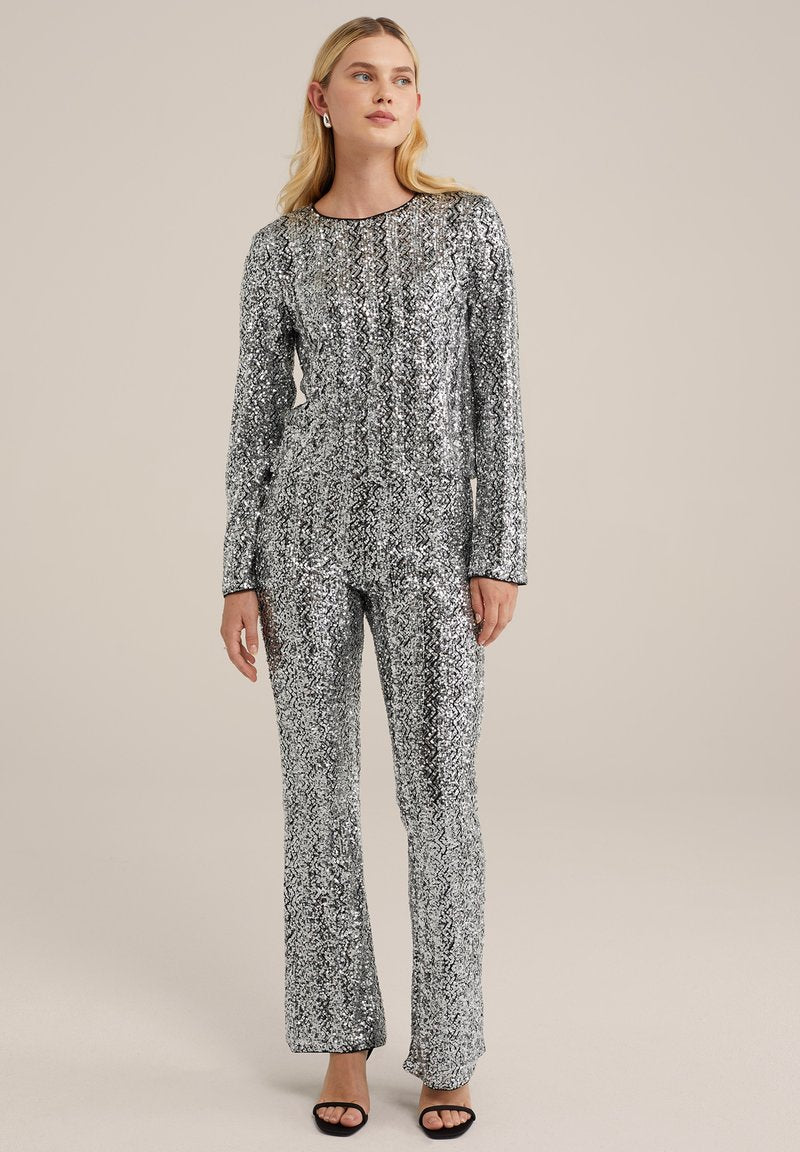 Silberner Pailletten-Overall mit langen Ärmeln und rundem Ausschnitt, der eine taillierte Taille und ausgestellte Hosen hat, kombiniert mit schwarzen hohen Sandalen.