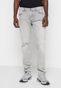 Mann trägt hellgraue, distressed Jeans mit Rissdetails an einem Knie, kombiniert mit einem schwarzen Shirt und grauen Sportschuhen.