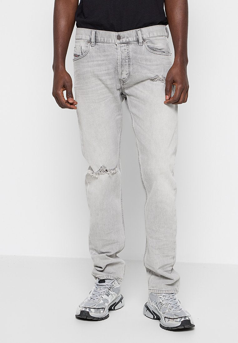 Mann trägt hellgraue, distressed Jeans mit Rissdetails an einem Knie, kombiniert mit einem schwarzen Shirt und grauen Sportschuhen.