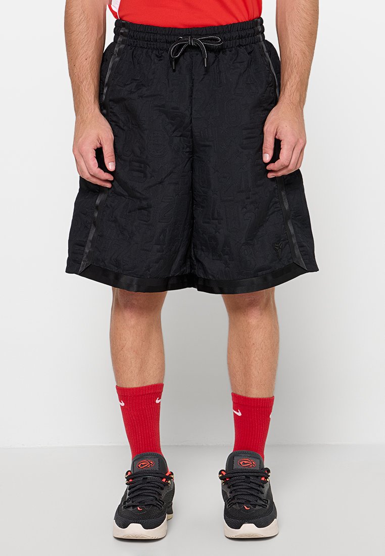 Mann trägt schwarze Sportshorts mit subtilen Mustern, rote Nike-Socken und schwarze Sportschuhe und steht vor einem schlichten Hintergrund.