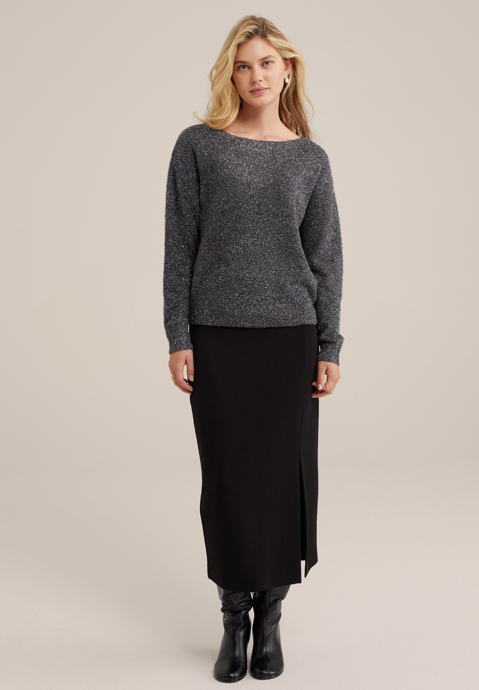 Grauer Strickpullover mit langen Ärmeln und weitem Halsausschnitt, kombiniert mit einem schwarzen Midirock mit seitlichem Schlitz und knielangen schwarzen Stiefeln.