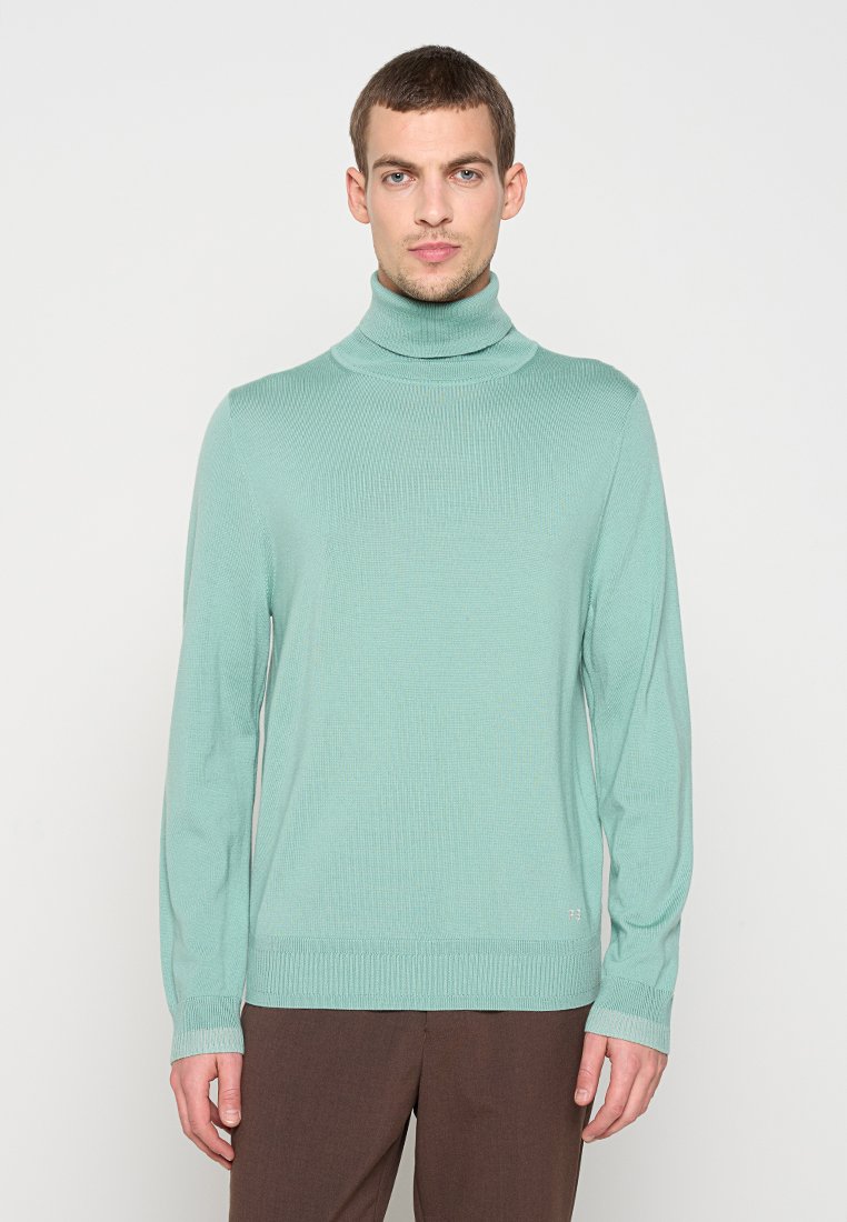 Hellgrüner Rollkragenpullover aus Rippstrick, mit langen Ärmeln und figurbetonter Silhouette, kombiniert mit braunen Hosen.