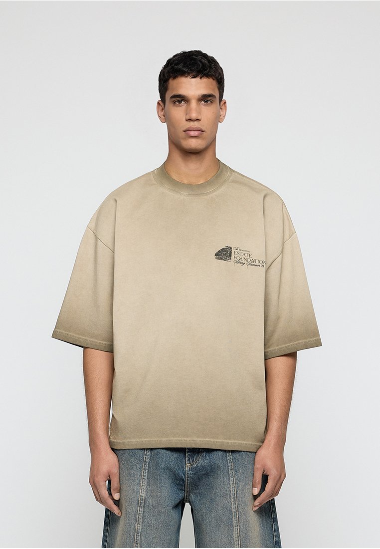 Junger Mann trägt ein übergroßes beiges T-Shirt mit kleinem schwarzem Logo auf der linken Brust und lockere blaue Jeans, steht vor einem schlichten Hintergrund.