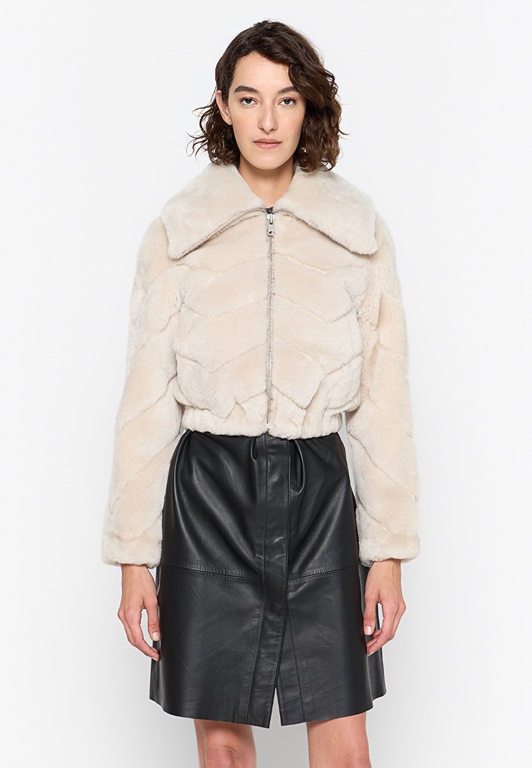 Kurze Jacke aus Kunstpelz in hellbeige mit Chevron-Muster, mit großem Kragen und Reißverschluss. Kombiniert mit einem schwarzen Lederrock.