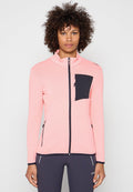 Person mit lockigem Haar, die eine pinke Reißverschlussjacke mit schwarzer Tasche und schwarze Hosen trägt, steht vor einem schlichten hellen Hintergrund.