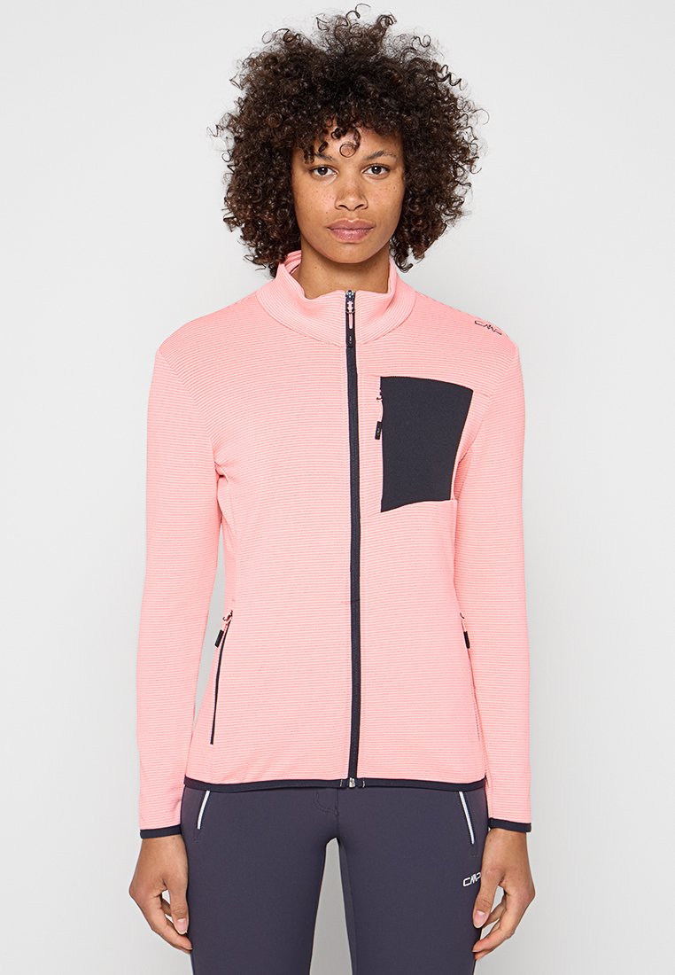 Person mit lockigem Haar, die eine pinke Reißverschlussjacke mit schwarzer Tasche und schwarze Hosen trägt, steht vor einem schlichten hellen Hintergrund.