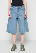 Person trägt hellblaue, weit geschnittene Jeansshorts, schwarzen Oberteil und grüne klobige Sneaker, steht vor einem schlichten hellen Hintergrund.