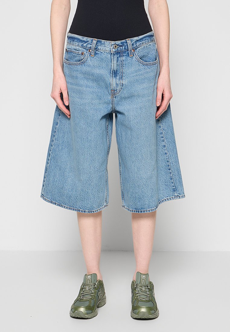 Person trägt hellblaue, weit geschnittene Jeansshorts, schwarzen Oberteil und grüne klobige Sneaker, steht vor einem schlichten hellen Hintergrund.