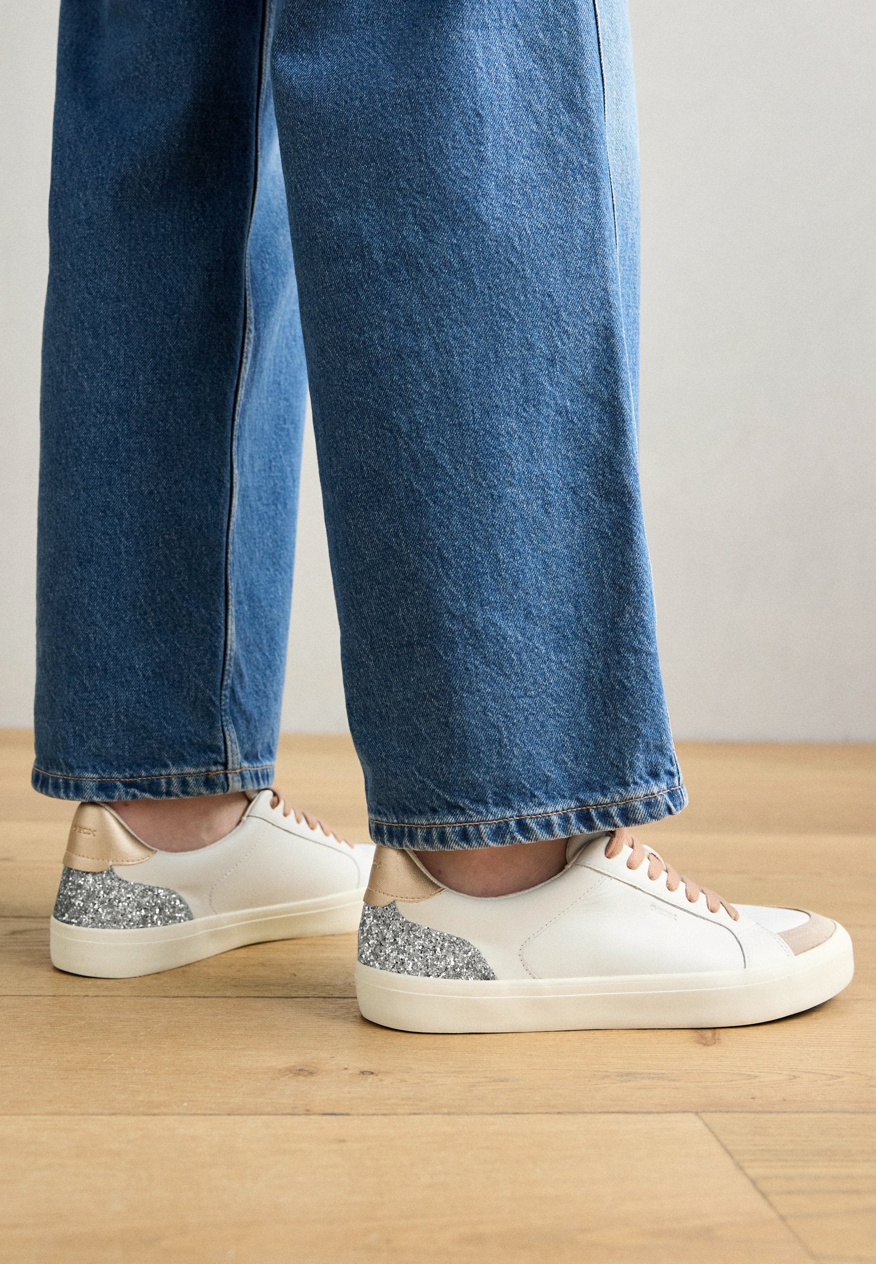Weiße Leder-Sneaker mit braunen Akzenten und silbernem Glitzer auf der Ferse, mit einer runden Zehenpartie und flachen Sohlen, kombiniert mit blauen Denim.