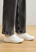 Weiße Sportschuhe mit Netzdetails, runder Zehe und flacher Gummisohle, getragen mit grauen weit geschnittenen Jeans auf einem Holzboden.