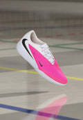 Weißer und pinker Nike Hallenfußballschuh mit schwarzem Swoosh-Logo, der über einem Hallenboden mit Sportplatzlinien schwebt.