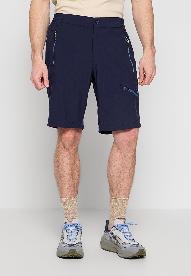 Mann trägt marineblaue Shorts mit Reißverschluss, beige Socken und graue Sneakers mit blauen Schnürsenkeln und steht vor einfachem Hintergrund.