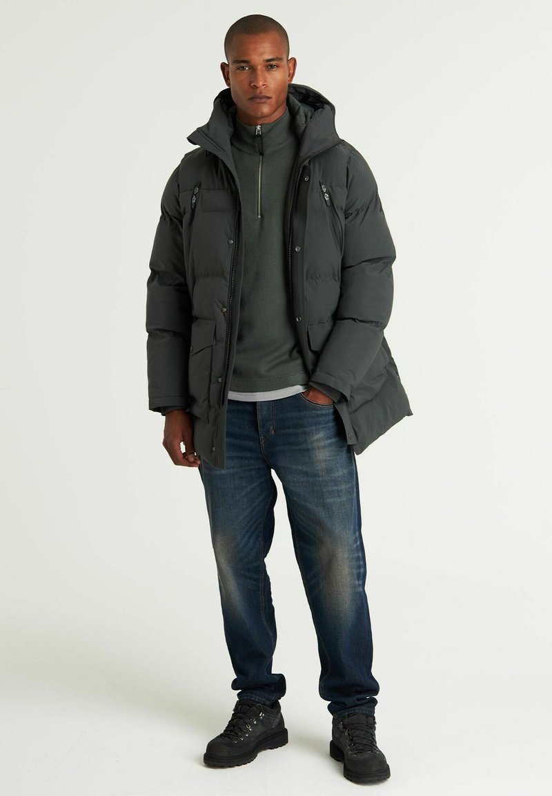 Dunkelgraue Pufferjacke mit Kapuze, Reißverschluss vorne und seitlichen Taschen, kombiniert mit einem grünen Pullover, blauen Jeans und schwarzen Stiefeln.