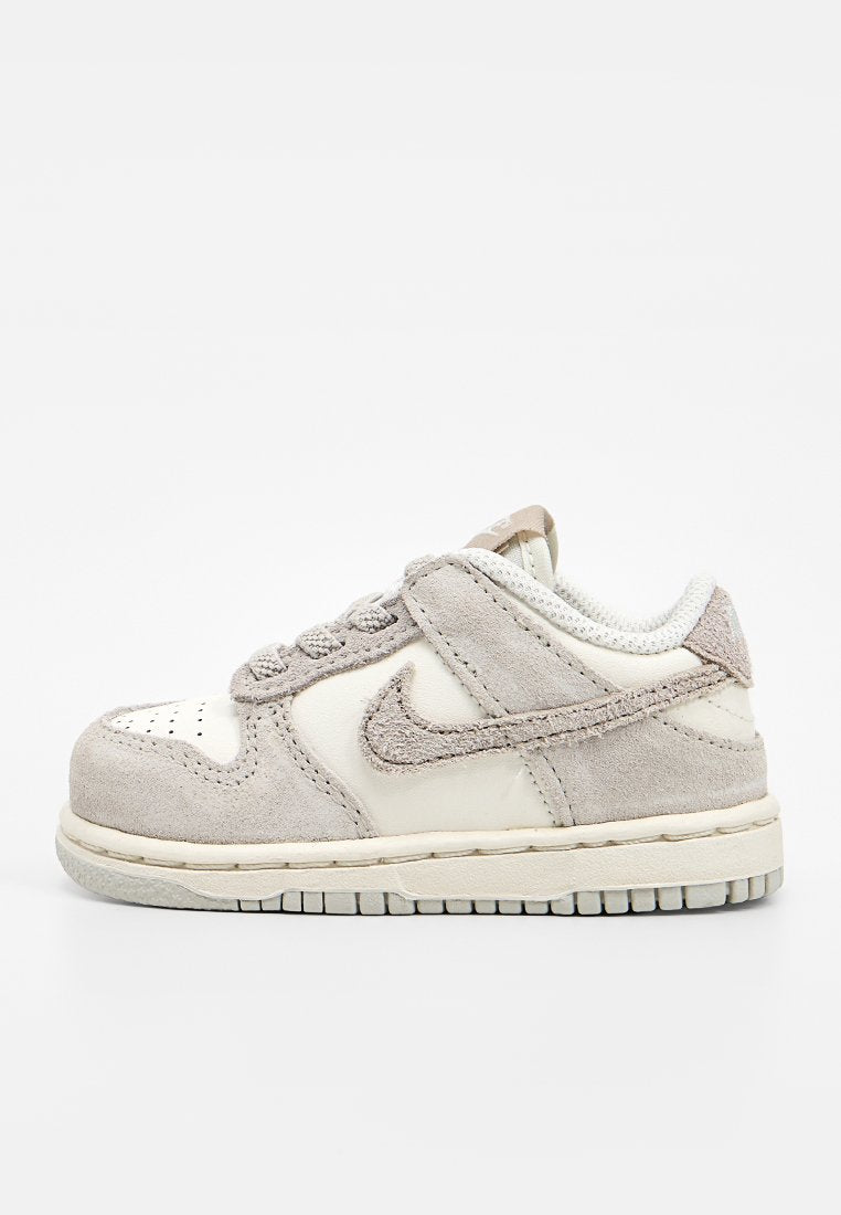 Hellbeiger und weißer Sneaker in Kleinkindgröße mit Wildleder-Overlays, Schnürsenkeln und Gummisohle, seitlich auf weißem Hintergrund dargestellt.
