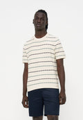 Junger Mann mit dunkler Haut und Dreadlocks trägt ein cremefarbenes kurzärmeliges Hemd mit dünnen horizontalen Streifen und dunkelblaue Shorts.
